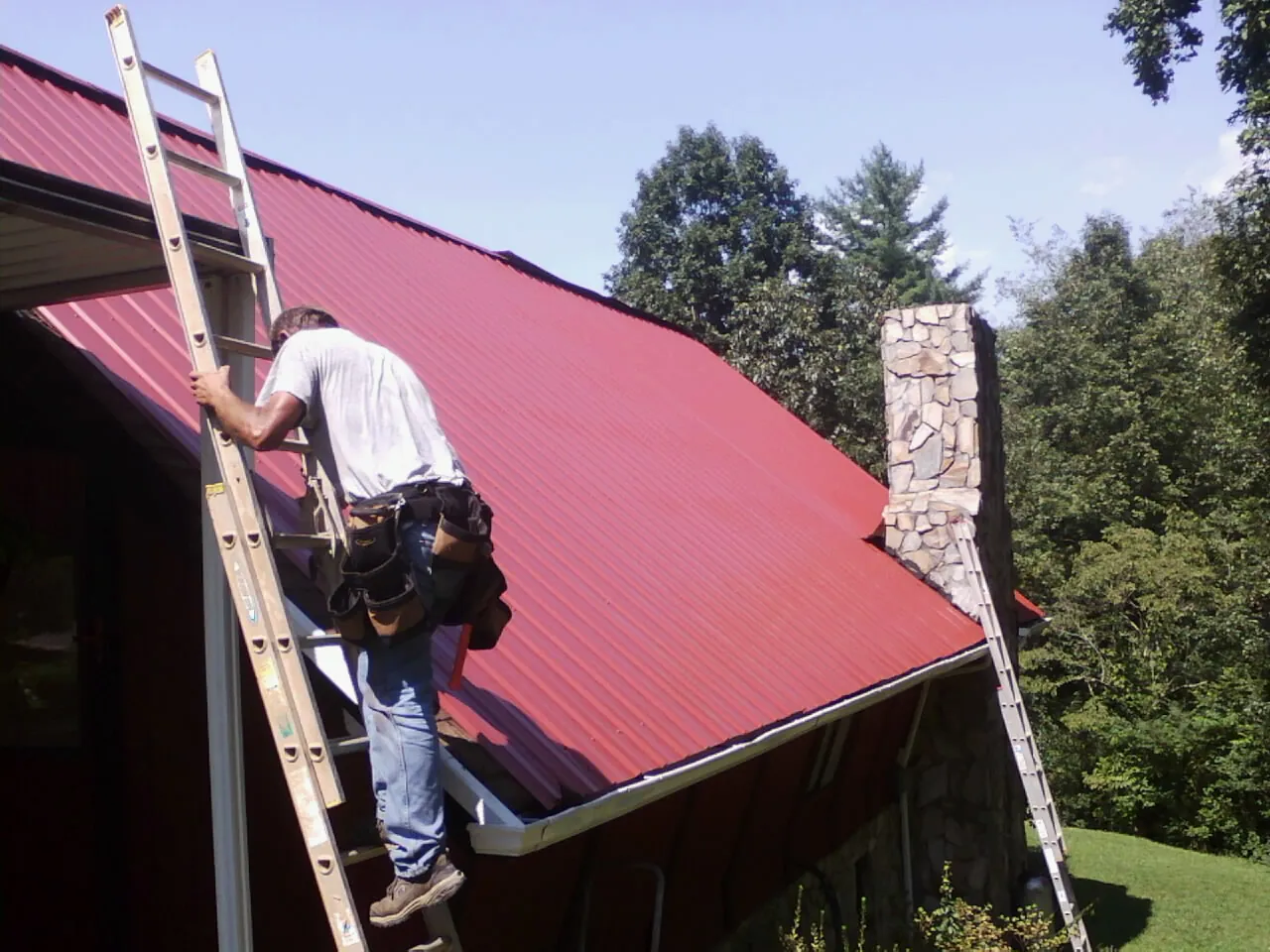 Roofer on a ladder beside a red metal roof and stone chimney — Soffit & Fascia Repair by Roofing Brothers in Palo
